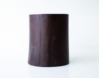 May include: A dark brown wooden stool shaped like a tree trunk. The stool has a smooth, rounded top and a slightly uneven base.