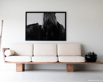 Può includere: Un divano bianco a tre cuscini con struttura e gambe in legno. Il divano si trova in un soggiorno minimalista con una fotografia in bianco e nero di una criniera di cavallo appesa al muro.