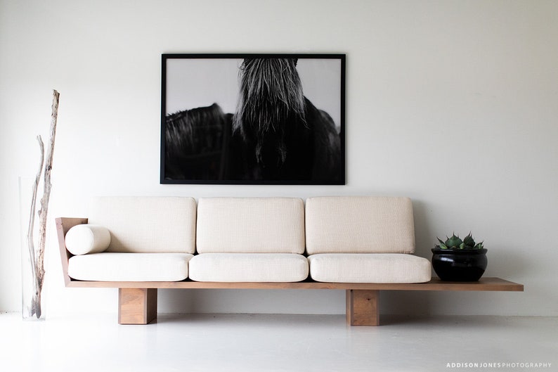 Pode incluir: Um sof&aacute; branco de tr&ecirc;s almofadas com estrutura e pernas de madeira. O sof&aacute; est&aacute; em uma sala de estar minimalista com uma fotografia em preto e branco de uma crina de cavalo pendurada na parede.