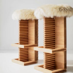 May include: Two wooden stools with a light-colored fur seat. The stools have a slatted design on the sides and a rectangular base. The wood has a natural finish, and the fur is a creamy white color. Modern furniture.