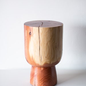 May include: A handcrafted wooden stool with a natural, organic design. The stool features a cylindrical top and a flared base, showcasing the wood's grain and color variations. The wood is a mix of light and reddish-brown tones.