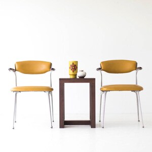 Puede incluir: Dos sillas de color amarillo mostaza con patas y brazos cromados, enfrentadas a trav&eacute;s de una mesa auxiliar cuadrada de madera oscura. Un jarr&oacute;n de cer&aacute;mica amarillo con un dise&ntilde;o floral rojo y naranja se encuentra sobre la mesa, junto con un peque&ntilde;o jarr&oacute;n de cer&aacute;mica blanco.