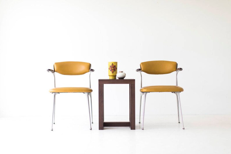 Puede incluir: Dos sillas de color amarillo mostaza con patas y brazos cromados, enfrentadas a trav&eacute;s de una mesa auxiliar cuadrada de madera oscura. Un jarr&oacute;n de cer&aacute;mica amarillo con un dise&ntilde;o floral rojo y naranja se encuentra sobre la mesa, junto con un peque&ntilde;o jarr&oacute;n de cer&aacute;mica blanco.