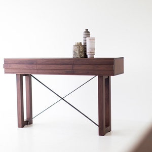 May include: A dark brown wooden console table with a black metal X-shaped base. The table has three drawers and is decorated with three ceramic vases.