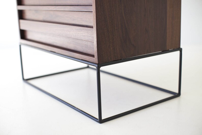 Pu&ograve; includere: Un mobile moderno in legno scuro con tre cassetti. Il mobile &egrave; sostenuto da una struttura in metallo nero geometrica. Il design &egrave; minimalista e la venatura del legno &egrave; visibile.