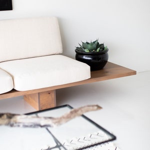 Pode incluir: Um sof&aacute; branco com uma moldura de madeira e um vaso preto com uma planta verde no topo da moldura. O sof&aacute; est&aacute; sentado em uma superf&iacute;cie branca.