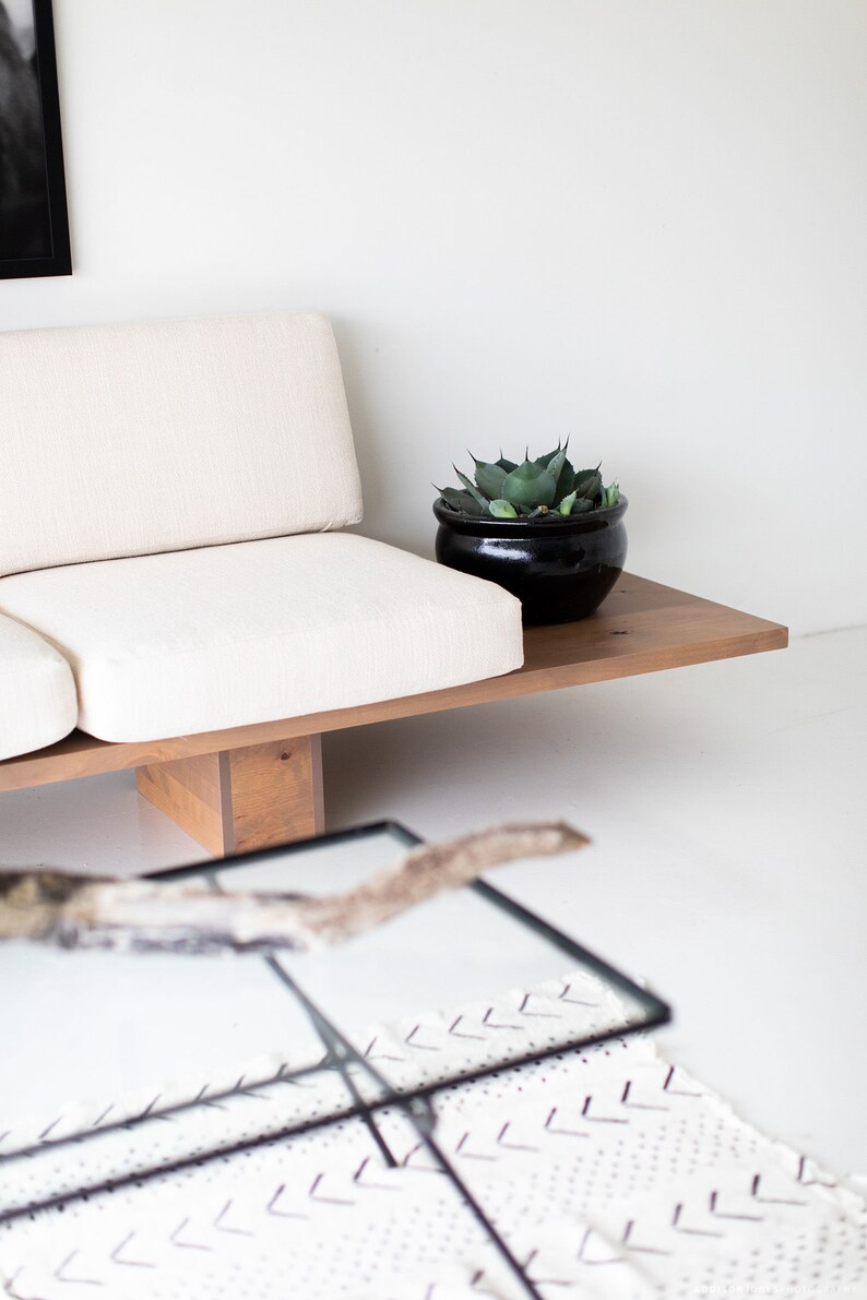 Pode incluir: Um sof&aacute; branco com uma moldura de madeira e um vaso preto com uma planta verde no topo da moldura. O sof&aacute; est&aacute; sentado em uma superf&iacute;cie branca.