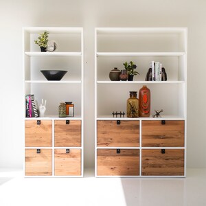 May include: Two white bookcases with open shelves and wooden drawers. The shelves are decorated with plants, vases, and books. The drawers are made of reclaimed wood.