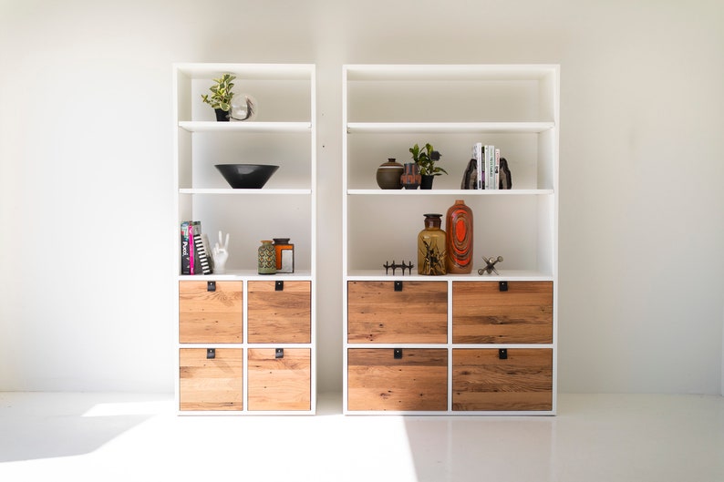 May include: Two white bookcases with open shelves and wooden drawers. The shelves are decorated with plants, vases, and books. The drawers are made of reclaimed wood.