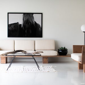 Pode incluir: Um sof&aacute; branco com uma moldura de madeira e uma fotografia em preto e branco de uma crina de cavalo pendurada na parede acima. Uma cadeira de madeira com uma almofada branca fica &agrave; direita do sof&aacute;. Uma mesa de centro de vidro com um galho de madeira no topo fica na frente do sof&aacute;. Um tapete branco est&aacute; no ch&atilde;o na frente do sof&aacute;.
