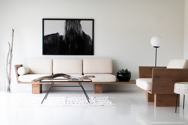 Pode incluir: Um sof&aacute; branco com uma moldura de madeira e uma fotografia em preto e branco de uma crina de cavalo pendurada na parede acima. Uma cadeira de madeira com uma almofada branca fica &agrave; direita do sof&aacute;. Uma mesa de centro de vidro com um galho de madeira no topo fica na frente do sof&aacute;. Um tapete branco est&aacute; no ch&atilde;o na frente do sof&aacute;.
