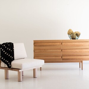 May include: A light wood dresser with six drawers and a white cushion chair with a black and white patterned throw blanket. The dresser has a simple design and is made of natural wood. The chair is a comfortable and stylish piece of furniture.