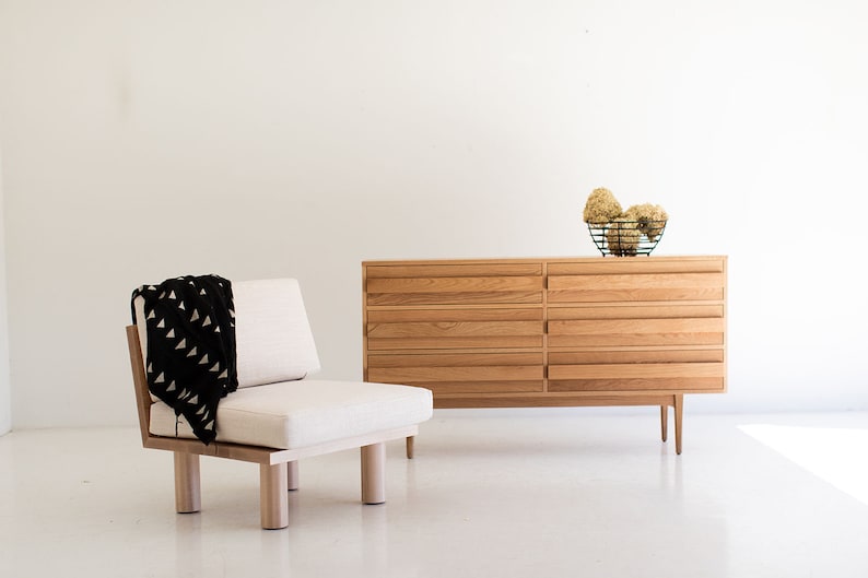 May include: A light wood dresser with six drawers and a white cushion chair with a black and white patterned throw blanket. The dresser has a simple design and is made of natural wood. The chair is a comfortable and stylish piece of furniture.