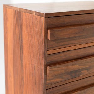 Pu&ograve; includere: Primo piano di un com&ograve; in legno con una ricca finitura marrone scuro. Il com&ograve; ha pi&ugrave; cassetti con scanalature orizzontali e un design pulito e moderno. La venatura del legno &egrave; visibile, aggiungendo consistenza.