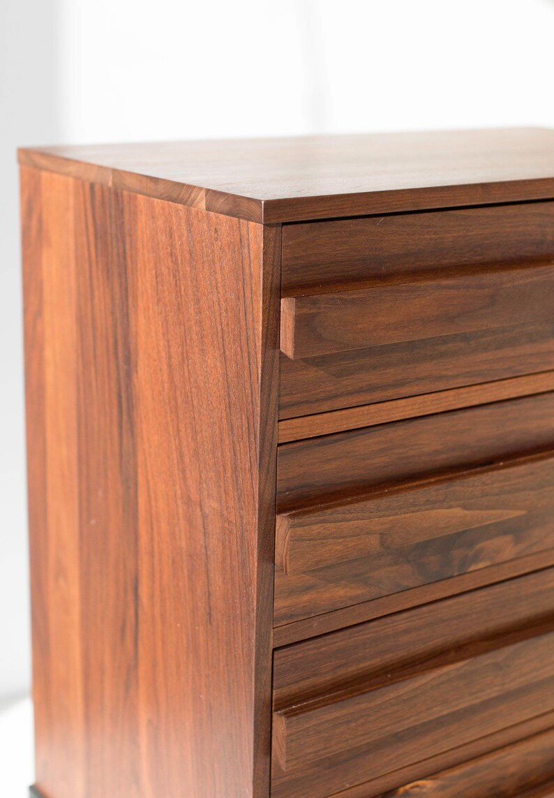 Pu&ograve; includere: Primo piano di un com&ograve; in legno con una ricca finitura marrone scuro. Il com&ograve; ha pi&ugrave; cassetti con scanalature orizzontali e un design pulito e moderno. La venatura del legno &egrave; visibile, aggiungendo consistenza.