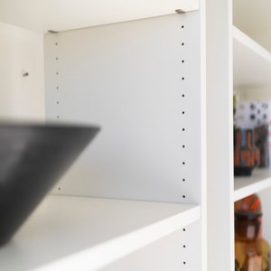 May include: White bookshelf with adjustable shelves. The shelves are made of white wood and have a smooth finish. The shelves are adjustable and can be moved to different heights.