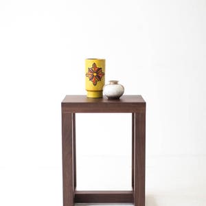 Puede incluir: Una mesa auxiliar de madera marr&oacute;n oscuro con una tapa cuadrada y un dise&ntilde;o de marco simple y abierto. Un jarr&oacute;n de cer&aacute;mica amarillo con un dise&ntilde;o floral rojo y naranja se encuentra sobre la mesa, junto con un jarr&oacute;n de cer&aacute;mica blanco m&aacute;s peque&ntilde;o.