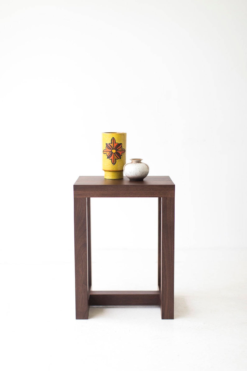 Puede incluir: Una mesa auxiliar de madera marr&oacute;n oscuro con una tapa cuadrada y un dise&ntilde;o de marco simple y abierto. Un jarr&oacute;n de cer&aacute;mica amarillo con un dise&ntilde;o floral rojo y naranja se encuentra sobre la mesa, junto con un jarr&oacute;n de cer&aacute;mica blanco m&aacute;s peque&ntilde;o.