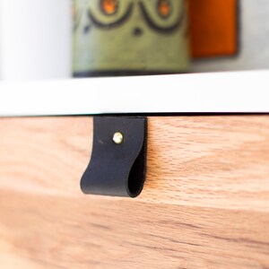 May include: A black leather drawer pull with a gold metal rivet. The pull is attached to a light wood drawer front.