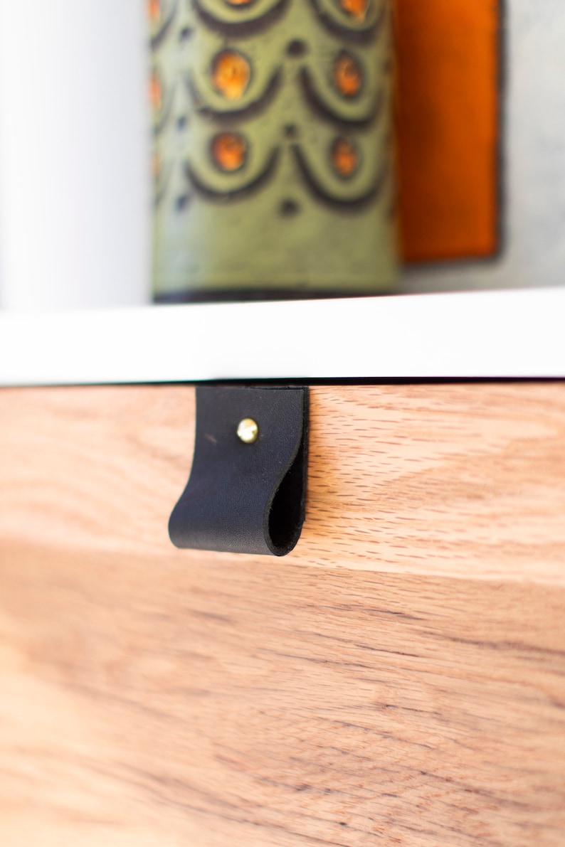 May include: A black leather drawer pull with a gold metal rivet. The pull is attached to a light wood drawer front.