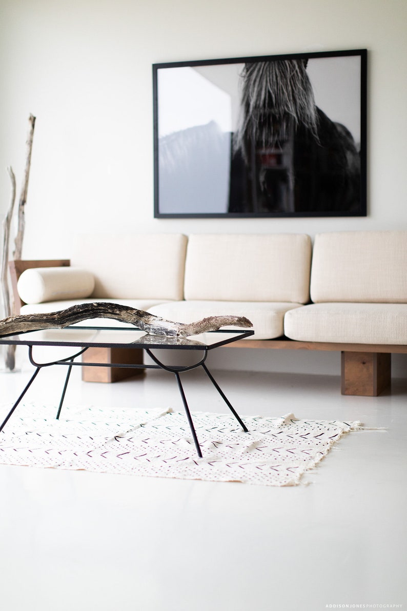 Pode incluir: Um sof&aacute; branco com uma fotografia de um cavalo emoldurada em preto acima. Uma mesa de centro de vidro com uma base de madeira fica na frente do sof&aacute;. Um ramo de madeira flutuante &eacute; colocado em cima da mesa. Um tapete branco com um padr&atilde;o preto est&aacute; no ch&atilde;o.