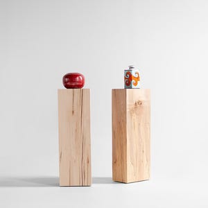May include: Two tall, rectangular light-colored wooden pedestals stand side by side. A smooth, round, dark red object rests on the left pedestal, while a small, square, white ceramic container with orange swirl patterns sits on the right pedestal.