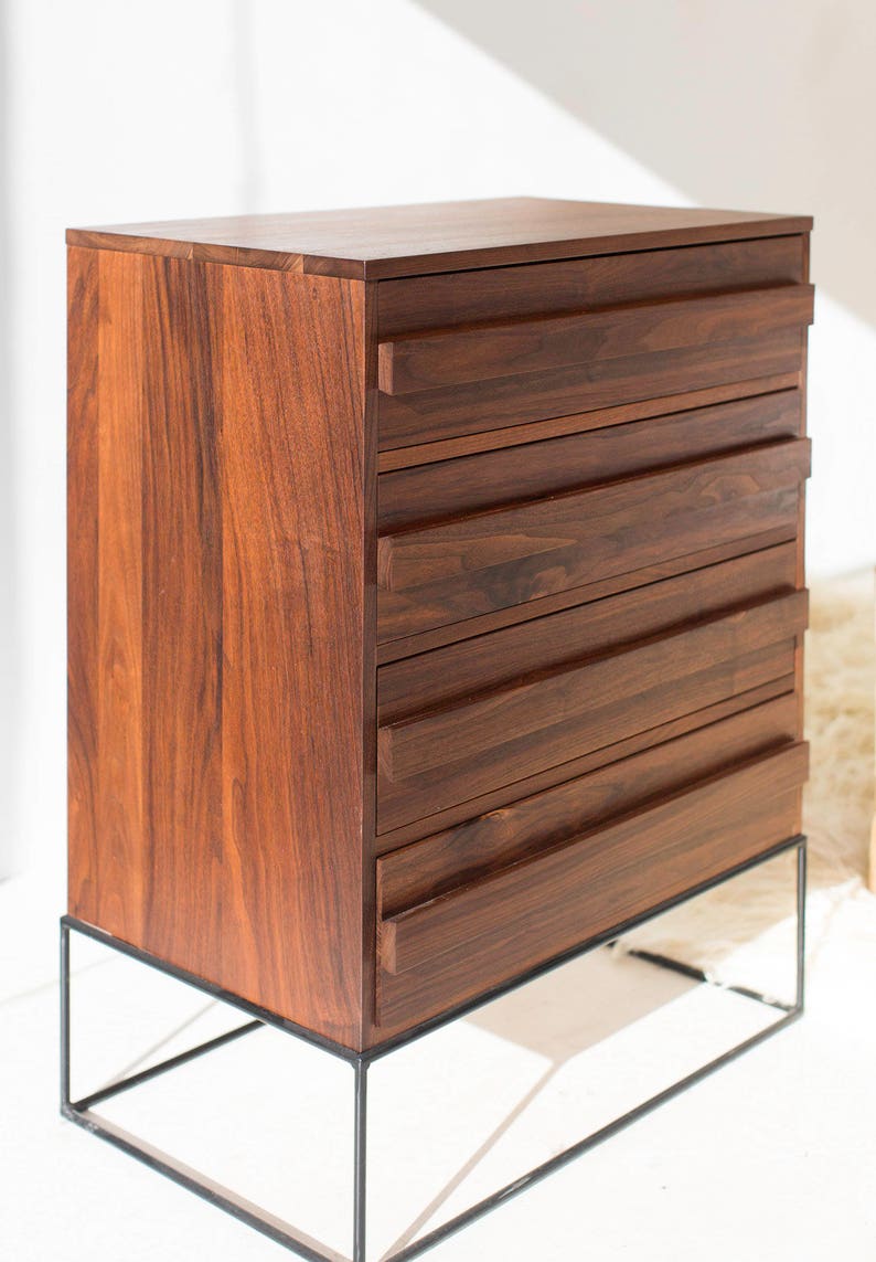 Pu&ograve; includere: Una cassettiera in legno scuro con cinque cassetti, posta su una struttura in metallo nero. La venatura del legno &egrave; visibile, mostrando un colore marrone caldo e ricco. I cassetti hanno un design minimalista. Una soluzione di archiviazione moderna.