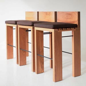 May include: Three wooden bar stools with brown cushions and a light wood backrest. The stools have a minimalist design with rectangular legs and a black metal footrest. The wood has a natural grain.