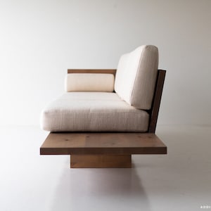 Pode incluir: Um sof&aacute; branco estofado com uma estrutura de madeira e uma base de plataforma baixa. O sof&aacute; tem um design simples e moderno.