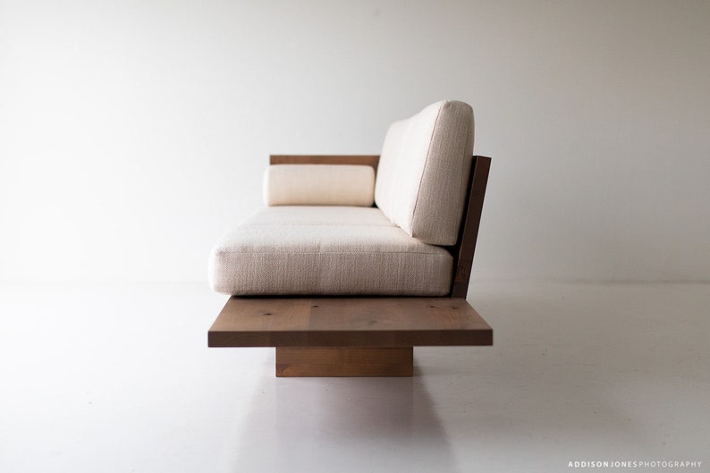 Pode incluir: Um sof&aacute; branco estofado com uma estrutura de madeira e uma base de plataforma baixa. O sof&aacute; tem um design simples e moderno.