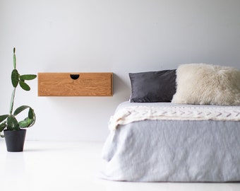 May include: A wooden floating nightstand with a drawer, mounted on a white wall above a bed with a gray and white blanket and two pillows. A small potted plant is on the left side of the image.