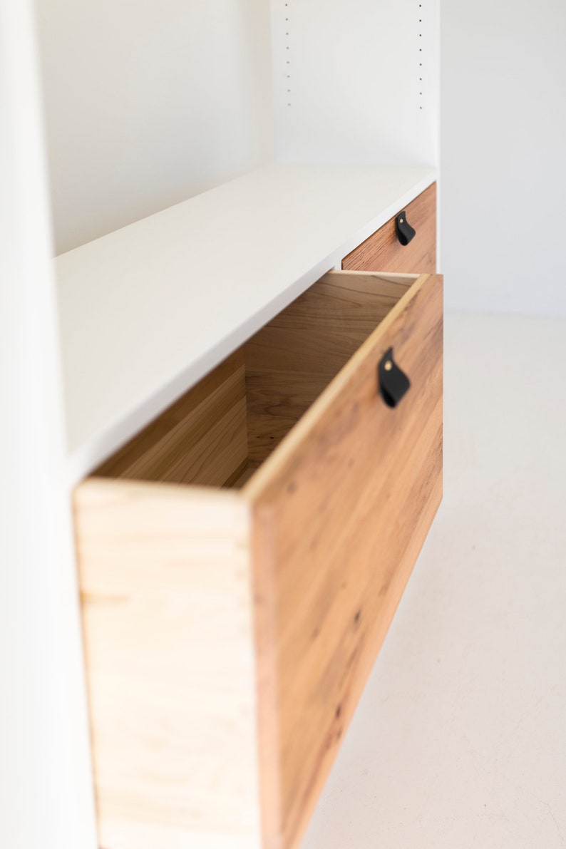 May include: A white cabinet with two wooden drawers. The drawers have black leather pulls. The top drawer is closed and the bottom drawer is open.