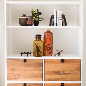 May include: White bookshelf with four wooden drawers. The top shelf has a small plant, two ceramic vases, and a set of bookends. The middle shelf has a glass bottle and a tall ceramic vase with orange and black stripes.