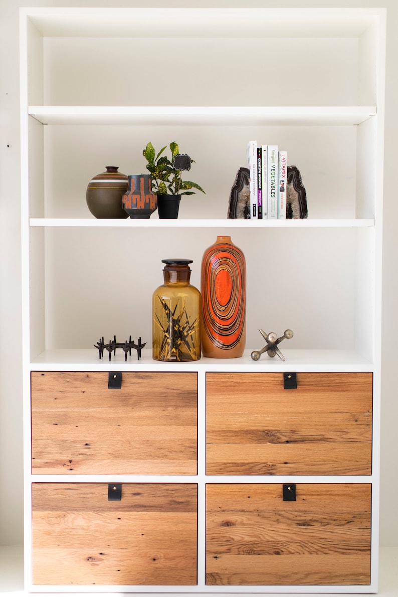 May include: White bookshelf with four wooden drawers. The top shelf has a small plant, two ceramic vases, and a set of bookends. The middle shelf has a glass bottle and a tall ceramic vase with orange and black stripes.