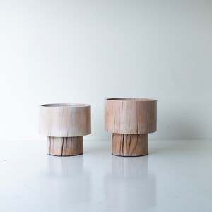 May include: Two wooden plant stands with a natural finish. The stands are made of two stacked pieces of wood, creating a unique and rustic look.