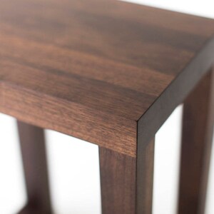 Puede incluir: Un taburete de madera marr&oacute;n oscuro con una parte superior cuadrada y un dise&ntilde;o de marco simple y abierto.