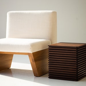 May include: A modern chair with a cream-colored upholstered seat and back, set on a light wood frame. Beside it is a dark brown, cube-shaped side table with a slatted design.