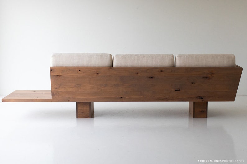 Pode incluir: Um sof&aacute; de madeira com um estofamento de tecido bege claro e uma prateleira de madeira por baixo. O sof&aacute; tem um design simples e moderno.