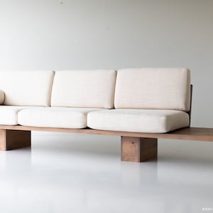 Pode incluir: Um sof&aacute; longo, baixo e de madeira com um assento de tecido branco e uma almofada combinando. O sof&aacute; tem um design simples e moderno e &eacute; suportado por quatro blocos de madeira.