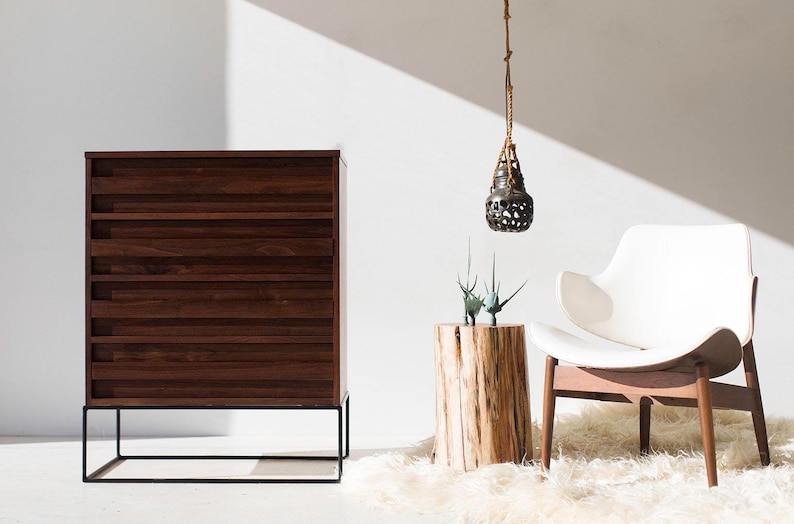 Pu&ograve; includere: Una scena d'interni moderna con un com&ograve; in legno scuro su una base in metallo nero, una sedia bianca con gambe in legno e un piccolo tavolino in legno. Una lampada a sospensione e un soffice tappeto bianco completano la scena.