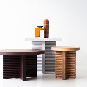 May include: Three round wooden tables with slatted bases. The table on the left is dark brown, the middle table is white, and the table on the right is light brown.