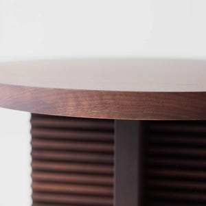 May include: A round, dark brown wooden table with a solid, rectangular base and a slatted, textured lower section. The table's top is smooth and polished, showcasing the natural wood grain.