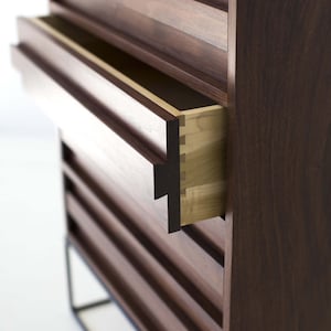 Pu&ograve; includere: Un moderno com&ograve; in legno con un cassetto parzialmente aperto, che rivela un interno di colore chiaro. Il com&ograve; &egrave; di colore marrone scuro con scanalature orizzontali e poggia su un telaio in metallo nero. L'incastro &egrave; visibile.