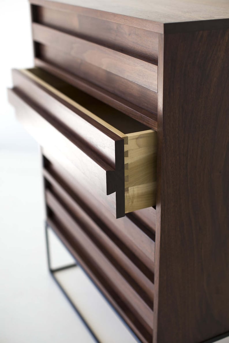 Pu&ograve; includere: Un moderno com&ograve; in legno con un cassetto parzialmente aperto, che rivela un interno di colore chiaro. Il com&ograve; &egrave; di colore marrone scuro con scanalature orizzontali e poggia su un telaio in metallo nero. L'incastro &egrave; visibile.