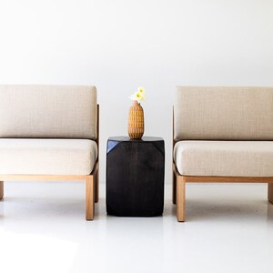 May include: Two modern armchairs with light beige cushions and wooden frames flank a dark, square side table. A decorative vase with yellow flowers sits on the table. The chairs and table are set against a white background.