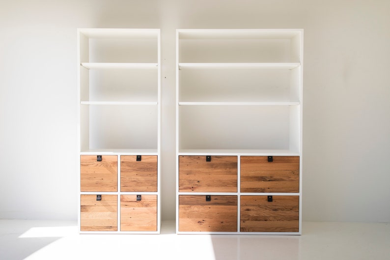 May include: Two white bookcases with open shelves and wooden drawers. The bookcases have a modern design and are perfect for storing books, decor, or other items.