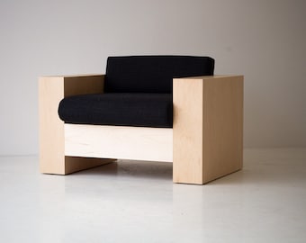 May include: Modern armchair with a black cushion and light wood frame. The chair has a minimalist design with a boxy shape and a low profile. The light wood has a natural finish, and the black cushion provides a contrast.