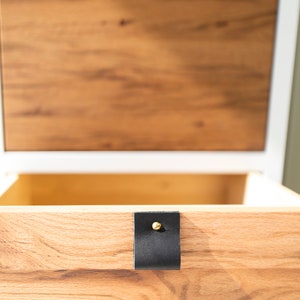 May include: A wooden drawer with a black leather pull and a gold metal knob. The drawer is open and the wood grain is visible.