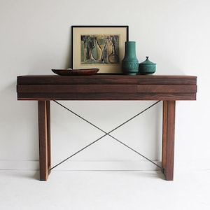 May include: A dark wood console table with two drawers and a metal X-shaped base. The table is decorated with a framed print, a green vase, and a green lidded jar.