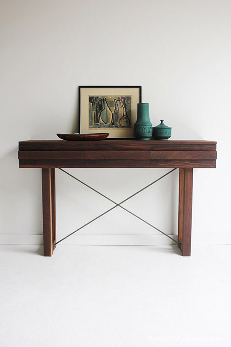 May include: A dark wood console table with two drawers and a metal X-shaped base. The table is decorated with a framed print, a green vase, and a green lidded jar.